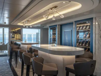Luxuriöse Fizz Champagne Bar an Bord der MSC World Europa. Elegante Bartresen, Barhocker, große Fenster mit Meerblick. Beleuchtete Weinregale und Marmorwände prägen das moderne Interieur.