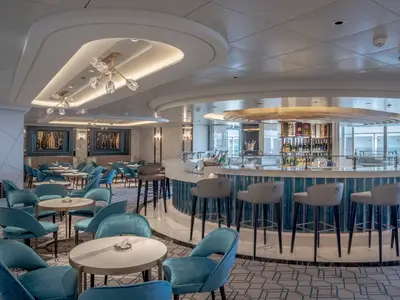Ansicht der eleganten Champagne Bar Fizz an Bord der MSC World Europa. Blaue Samtsessel, helle Tische und eine runde Bar mit Champagnerflaschen. Tageslicht, Meerblick.