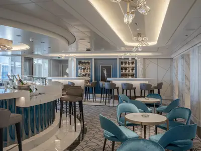 Blick in die Champagne Bar Fizz an Bord der MSC World Europa. Ein geschwungener Bartresen mit blauen Fliesen und Marmor, hohe Barhocker und blaue Samtsessel. Gekühlter Champagner ist sichtbar, ergänzt durch moderne Lichtinstallationen und Marmorwände.