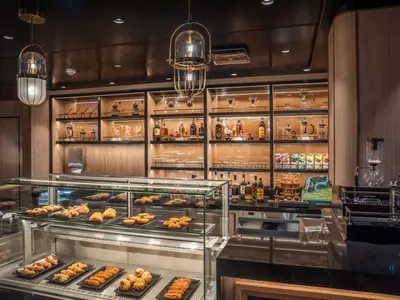 Eine elegante Kaffee-Bar an Bord der MSC World Europa. Die Theke zeigt frisches Gebäck in einer beleuchteten Glasvitrine, während dahinter Spirituosen und Kaffeebereiter auf Holzregalen präsentiert werden.