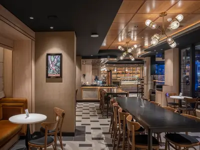 Das helle Coffee Emporium an Bord der MSC World Europa mit Holzelementen, dunklen Tischen und stylischen Deckenleuchten. Im Hintergrund eine Bar mit Patisserie. Eine Sitzecke mit einer orangefarbenen Bank ist links im Bild.