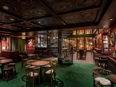Ein gemuetlicher Pub an Bord der MSC World Europa. Rote Waende mit Bildern, gruener Teppich, dunkle Holzmoebel, eine Wendeltreppe und die Brauerei sind zu sehen.