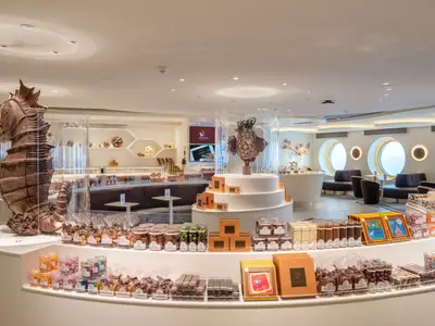 Innenansicht des Jean-Philippe Chocolat & Café an Bord der MSC World Europa. Im Vordergrund sind Auslagen mit Pralinen und Süßigkeiten zu sehen, sowie große Schokoladen-Skulpturen eines Seepferdchens und eines Fisches. Im Hintergrund gibt es Sitzbereiche und Bullaugenfenster.