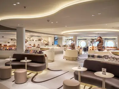 Moderner Schokoladenladen an Bord der MSC World Europa. Mit Pralinen gefüllte Auslagen, elegante Sitzbereiche und große Schokoladenskulpturen, darunter ein Seepferdchen, sind zu sehen.