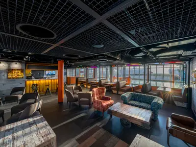 Die 'The Box Bar' an Bord der MSC World Europa zeigt eine moderne Bar mit Betonelementen, Gitterdecke und Fensterblick. Verschiedene Sitzgelegenheiten wie Sessel und Bänke sind vorhanden. An der Wand hängen Skateboards als Dekoration.