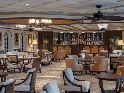 Die elegante Bar-Lounge 'Raj Polo Tea House' an Bord der MSC World Europa mit bequemen Sitzgelegenheiten, Holztischen und einer Bar im Hintergrund.