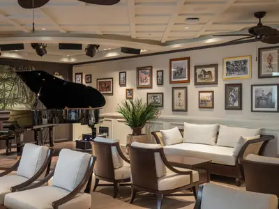 Elegante Lounge im Raj Polo Tea House an Bord der MSC World Europa mit einem Grand Piano auf einer Bühne. Viele gerahmte Bilder schmücken die Wände über bequemen Sesseln und Sofas.