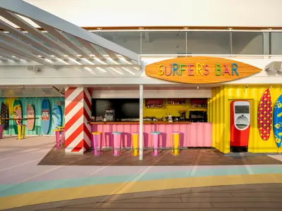 Die lebhafte Surfer's Bar an Bord der MSC World Europa zeigt eine pinke Theke, bunte Hocker, gelbe Wände und dekorative Surfbretter. Eine Säule in Rot-Weiss-Streifen fällt auf.