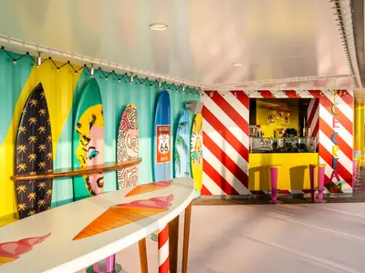 Eine lebhafte Surfer's Bar an Bord der MSC World Europa. Die Wände sind mit Streifen und bunten Pop-Art-Surfbrettern dekoriert, darunter eine Eiscreme-Bar mit leuchtend gelbem Tresen und pinken Hockern.