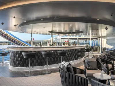 Detailansicht einer eleganten Aussenbar mit geschwungenem Tresen, Barhockern und bequemen Lounge-Sesseln auf dem Deck der MSC World Europa. Im Hintergrund ist das Meer zu sehen.