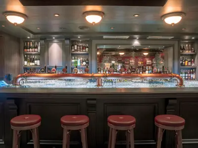 Bar 'Masters of the Sea' auf der MSC World Europa. Kupfere Zapfanlage, Spirituosenregale, Gläser und rote Barhocker. Hinten ein Spiegel mit Schiffs-Motiv.