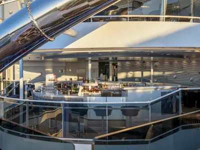 Die moderne Zen Bar an Bord der MSC World Europa. Geschwungene Bar mit Getränken, Barhockern. Eine glänzende Wasserrutsche ist links sichtbar. Maritime Wanddekoration mit Muscheln.