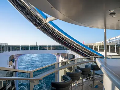 Die Zen Bar der MSC World Europa. Ein Außendeck mit Barhockern, Meerblick und einer grossen, glänzenden Röhrenrutsche. Im Hintergrund ein Deck mit Sonnenliegen.