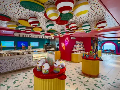 Innenansicht des bunten Süßigkeitenladens 'Sweet Temptations' an Bord der MSC World Europa mit Eistheke, Süßigkeiten und Spielzeug. Rundfenster geben den Blick auf das Meer frei.