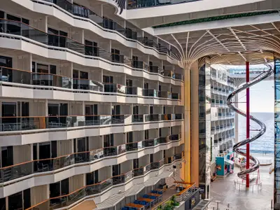 Innenansicht der MSC World Europa zeigt die World Promenade. Links viele Balkonkabinen, rechts eine hohe Spiralrutsche mit Meerblick. Moderne Architektur und einige Gäste.