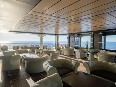 Die helle The Lanai Bar an Bord der MSC World Europa mit bequemen, cremefarbenen Sesseln, kleinen Tischen und dunklem Holzboden. Grosse Fenster bieten einen weiten Blick auf das blaue Meer und den Horizont.