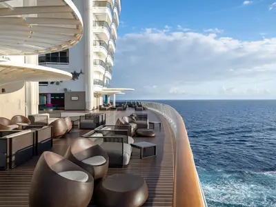 Die Lanai Bar an Bord der MSC World Europa. Das Außendeck ist mit modernen, braunen Sitzgruppen ausgestattet. Rechts erstreckt sich das weite, blaue Meer unter einem leicht bewölkten Himmel.