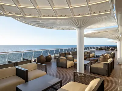Outdoor-Lounge auf der MSC World Europa: Moderne Sitzgruppen und Tische unter einer geschwungenen, weißen Überdachung mit Meerblick. Ohne Personen.