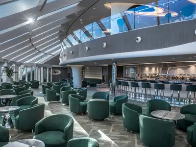 Die Top Sail Lounge an Bord der MSC World Europa mit modernen grünen Sesseln, glänzenden Tischen und einer eleganten Bar mit Hockern. Grosse Panoramafenster und Oberlichter bringen viel Tageslicht und Meerblick in den Raum.