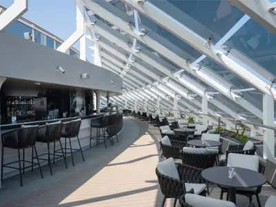 Blick auf die moderne Bar des MSC Yacht Club Grill & Bar an Bord der MSC World Europa. Die langgezogene Bar mit Hockern und bequemen Lounge-Sesseln unter einem Glasdach.