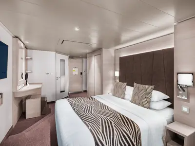 Ansicht einer modernen barrierefreien Deluxe-Balkonkabine an Bord der MSC World Europa mit Doppelbett, Sitzgelegenheiten und TV. Die Kabine ist hell und funktional eingerichtet.