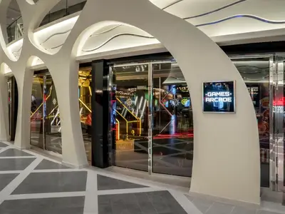 Eingang der Games Arcade auf der MSC World Europa. Helle, geschwungene Architektur und Glastüren. Bunte Neonlichter von Spielen sichtbar. Leuchtschild zeigt 'Games Arcade'.