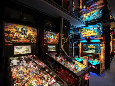 Ein moderner Spielsalon mit Flipperautomaten und Arcade-Spielen wie Wild West Shootout an Bord der MSC World Europa. Die leuchtenden Bildschirme der Automaten erhellen den dunklen Raum.