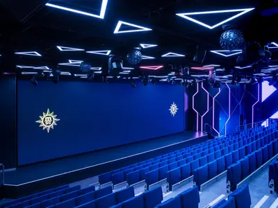 Die Luna Park Arena an Bord der MSC World Europa. Blaue LED-Bildschirme mit MSC-Logo und geometrische Neonlichter schmücken die Bühne und Wände. Davor stehen unbesetzte blaue Sitze in Reihen.
