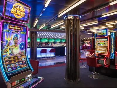 Das stilvolle Signature Casino an Bord der MSC World Europa. Leuchtende Spielautomaten im Vordergrund, eine moderne Bar mit Flaschen im Hintergrund und bequeme Barhocker.