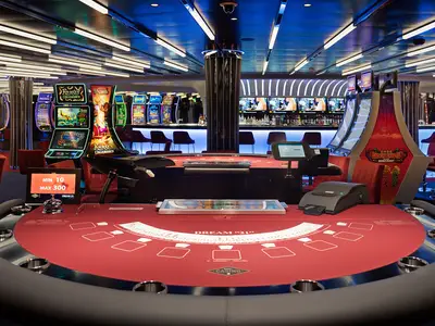 Ein modernes Casino an Bord der MSC World Europa, mit einem roten Blackjack-Tisch im Vordergrund, flankiert von leuchtenden Spielautomaten und einer schicken Bar im Hintergrund.