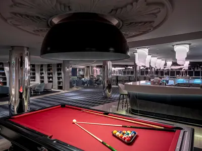 Die moderne Panorama Lounge an Bord der MSC World Europa mit einem roten Billardtisch im Vordergrund. Im Hintergrund erstreckt sich eine beleuchtete Bar, bequeme Sitzbereiche und Regale mit Dekorationen unter stilvollen Leuchten.