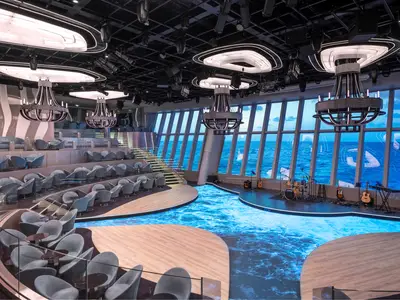 Die geräumige Panorama Lounge an Bord der MSC World Europa bietet eine Bühne mit Musikinstrumenten, elegante Sitzbereiche auf mehreren Ebenen und riesige Fenster mit Blick auf das blaue Meer. Eine beeindruckende Lichtinstallation ziert die Decke.