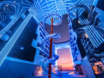 MSC World Europa: Die World Promenade mit blauer Beleuchtung und der hohen, transparenten Spiralrutsche. Im Hintergrund ein orange-rosafarbener Sonnenuntergang.