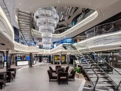 Panoramablick auf die moderne, mehrstöckige World Galleria an Bord der MSC World Europa. Ein glitzernder Kronleuchter dominiert den Raum, umgeben von geschwungenen Balustraden, eleganten Treppen und Sitzbereichen.