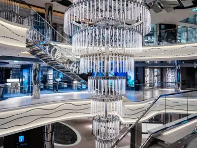 Blick in das futuristische, mehrstöckige Atrium an Bord der MSC World Europa. Ein riesiger, zylindrischer Kronleuchter dominiert den Raum, umgeben von eleganten geschwungenen Treppen und Geschäften mit Glasfronten.