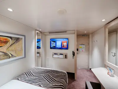 Eine moderne Innenkabine an Bord der MSC World Europa mit einem Einzelbett, bedeckt mit einem Zebra-Muster. Ein großer Fernseher und ein Kunstwerk schmücken die Wände. Eine Tür zum Flur ist sichtbar.