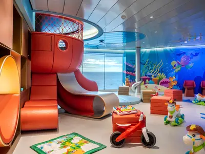 Ein geräumiger Baby Club an Bord der MSC World Europa mit einem großen roten Spielturm, Rutsche, weichen Sitzgelegenheiten und Spielzeug. Eine Wand ist mit einem farbenfrohen Unterwasser-Wandbild dekoriert, während große Fenster einen weiten Blick auf das offene Meer bieten.