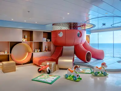 Kinderbereich Baby Club Chicco an Bord der MSC World Europa mit roten Rutschen, Schaukelpferden und einem runden Lesesessel. Große Fenster bieten Meerblick.