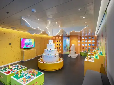 Bunter Kinderbereich an Bord der MSC World Europa mit Lego-Duplo-Spieltischen und großer Torte in der Mitte. Gelbe Wände und ein TV sind sichtbar.