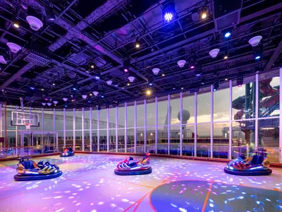 Lebhafter Indoor-Sportplex der MSC World Europa. Bunte Bumper-Cars stehen auf lila-blau beleuchtetem Spielfeld. Fenster zeigen Wasserrutschen und das Meer bei Dämmerung. Ohne Personen.