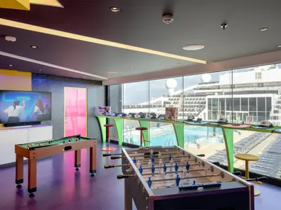 Im Young Club der MSC World Europa: Billard, Kicker, grosser TV-Bildschirm und eine pinke Tür. Durch die Fenster sind das Pooldeck und ein angedocktes Schiff zu sehen.