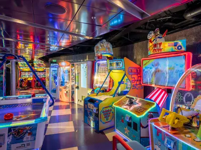 Eine bunte Spielhalle auf der MSC World Europa mit einer Vielzahl leuchtender Arcade-Automaten, darunter Airhockey, Rennspiele und Klauengreifer. Die Decke reflektiert die farbigen Lichter der Spiele.