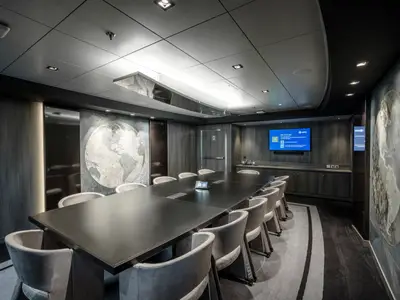 Ein modernes Business Center an Bord der MSC World Europa. Der lange Konferenztisch ist von bequemen Stühlen umgeben. An der Wand hängt ein Weltkarten-Kunstwerk, und ein großer Bildschirm zeigt MSC-Informationen. Die Beleuchtung ist hell und das Dekor elegant.