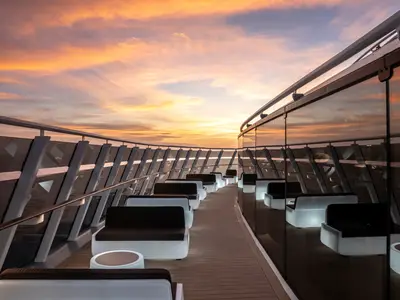 Elegante Aussen-Lounge der MSC World Europa bei Sonnenuntergang. Moderne Lounge-Möbel mit leuchtenden Sockeln und eine Glasfassade spiegeln den farbenprächtigen Himmel wider. Ohne Personen.