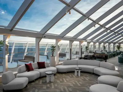 Eine moderne und helle Lounge an Bord der MSC World Europa. Große Fenster bieten Meerblick, ein Glasdach lässt viel Tageslicht herein. Bequeme Sofas, Sessel und ein Flügel prägen den Raum.