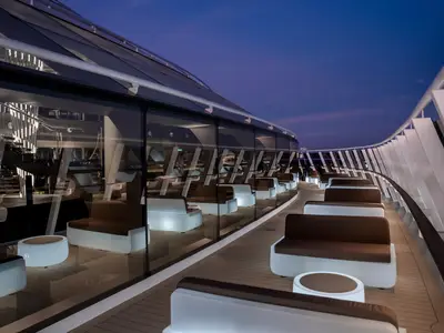 Abendliche Top Sail Lounge auf der MSC World Europa. Moderne Sitzmöbel auf Holzdeck. Glaswand reflektiert Innenraum. Geschwungene Geländer unter blauem Himmel.