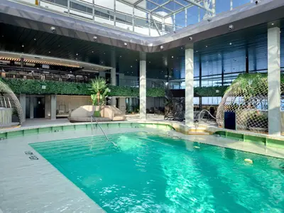 Ein grosser, lichtdurchfluteter Indoor-Poolbereich des Botanic Garden Pool an Bord der MSC World Europa mit üppiger Vegetation, bequemen Lounge-Möbeln und einem Oberdeck.
