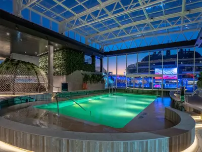 MSC World Europa: Überdachter Botanic Garden Pool. Grün beleuchteter Pool, Whirlpool, Pflanzen, Netzkuppel. Blick auf Oberdeck bei Blaue Stunde. Ohne Personen.