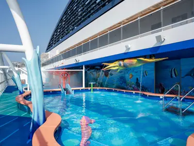 Farbenfroher Kinder-Aquapark an Bord der MSC World Europa. Flacher Pool mit Wasserspielen, Rutschen und Meeres-Wandmalerei. Großer Tintenfisch spritzt Wasser. Bei hellem Tageslicht auf dem Deck.