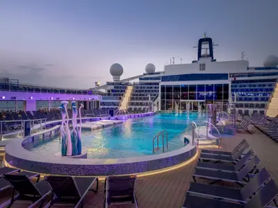 Abendliche Stimmung am La Plage Pool an Bord der MSC World Europa. Der große Pool und die Liegestühle sind in blau-violettes Licht getaucht. Eine moderne Skulptur und eine beleuchtete Treppe sind zentral.